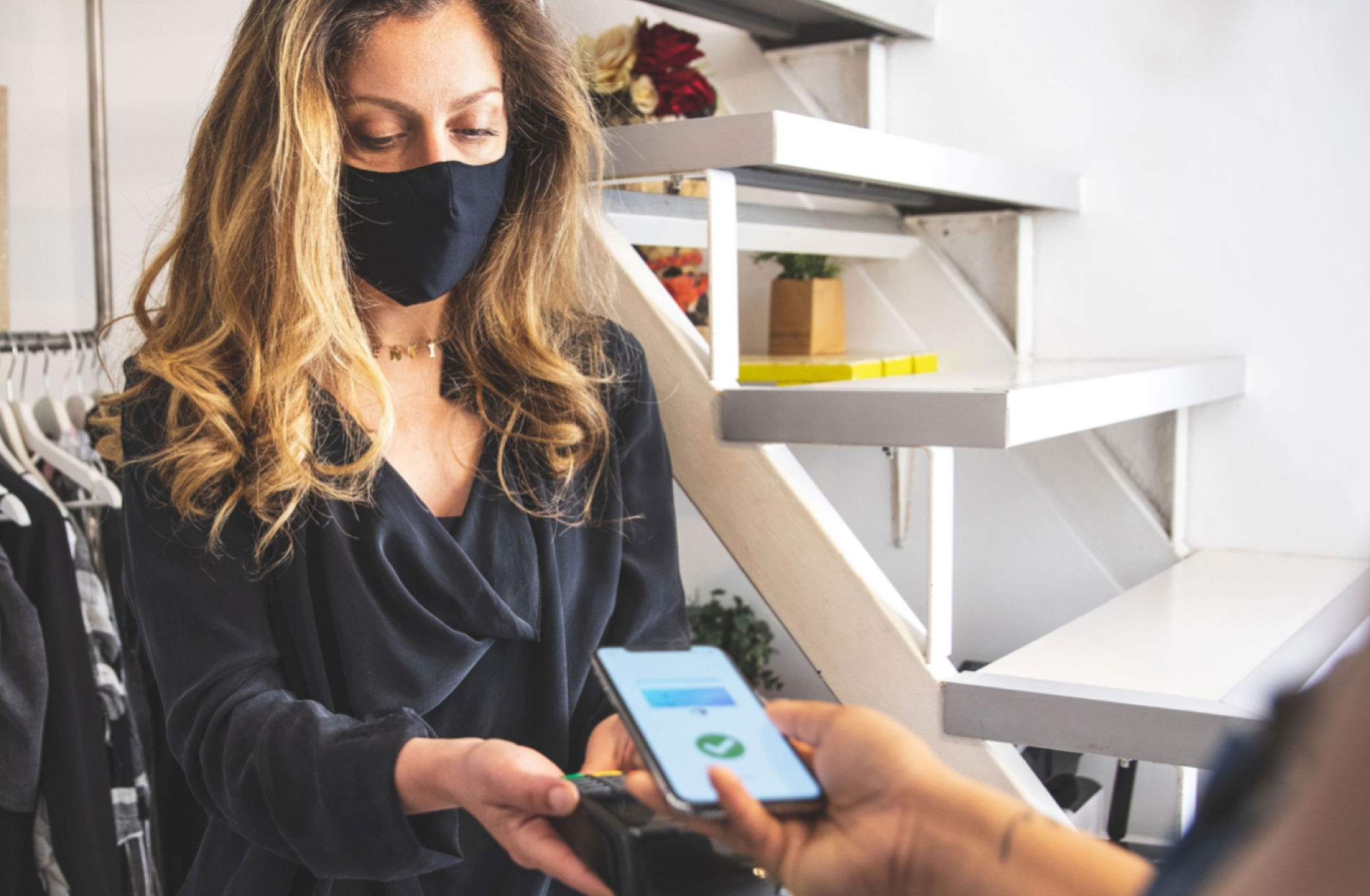 Woman in mask helps shopper to pay for retail purchases on the floor via her smart phone.